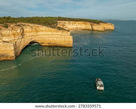 Drone view of Benagil Cave cliffs in Algarve, Portugal, with a tourist tour boat on Atlantic water, limestone sea caves, and rugged coast.