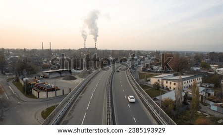 Drone view of the bridge passing through the city. Different cars pass by, from cars to trucks. Free lane. Against the background of a city at sunset in smog. The chimneys of the factory are smoking