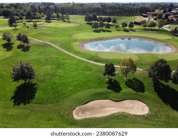 Drone view of green grassy golf course with small pond water reflecting blue cloudy sky sandy terrain patch walking path across grass and trees with buildings in sunny daylight - Powered by Shutterstock