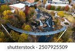 Drone view of Liberty Bridge over the Reedy River at Falls Park, Downtown Greenville, SC. Vibrant fall foliage and urban architecture frame the cascading waterfalls in this scenic landscape