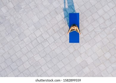 Drone View Of Quiet Healthy Person Comfortably Sitting On Blue Yoga Mat In Lotus Pose And Recreating In Daylight On Stone Floor