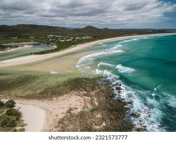 Drone view of white sandy beach with river inlet and rocky shore. Coastal township, green  lush mountains, and a bridge across the river. People are swimming, surfing and playing in the sea. - Powered by Shutterstock