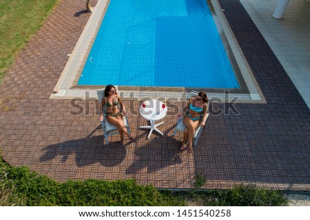 Drone view of young women sunbathing, drinking cocktails and relaxing by swimming pool in summer