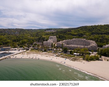 drone's eye view of the beach, sea, boat dock, modern hotel, and forest in the distance. Perfect for vacation, travel, and leisure lovers seeking a serene and picturesque escape