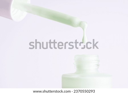 Drop falls from a pipette into a cosmetic bottle on white background, macro. Pastel green face cream, makeup primer, color correcting cosmetic product, serum