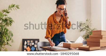 Drop shipping business owner talking on mobile phone and taking order. Female entrepreneur working at home office confirming the order on phone.