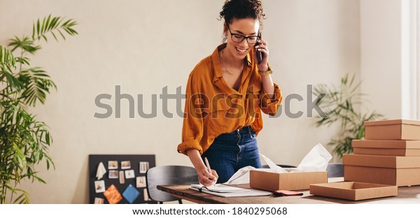 Drop shipping business owner talking on mobile\
phone and taking order. Female entrepreneur working at home office\
confirming the order on\
phone.