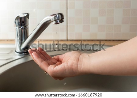 A drop of water drips from faucet in human palm above round metal kitchen sink