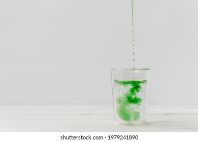 Drops Of Liquid Chlorophyll Falling Down In A Glass Cup With Water. Light White Background With Copy Space