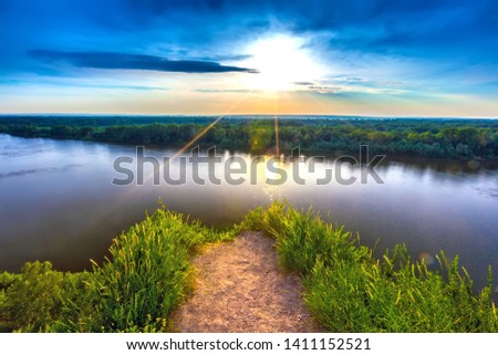 Drowned gravel dangerous path before a cliff overgrown green grass, rise the White River during sunset under a clear blue sky against the setting sun. Rock Hanging Stone, Ufa, Bashkortostan, Russia.