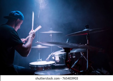 Drummer in a cap and headphones plays drums at a concert under blue light in a smoke