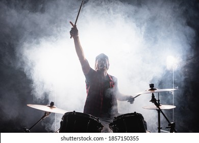 Drummer with hand in air, holding drumsticks while sitting at drum kit with smoke on black