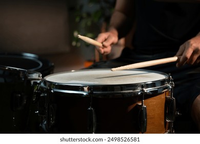 A drummer plays a snare drum in a dimly lit studio setting. The drummer is wearing a black shirt and uses a wooden drumstick to play the drum. The snare drum is a standard size, wooden drum with - Powered by Shutterstock