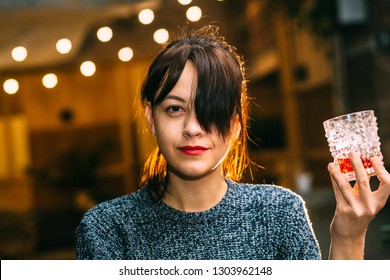 Drunk Brunette Student Woman Alone In Happy Smiling Face Expression Holding And Looking Thoughtful To Scotch Whiskey Glass At Bar Or Pub In Alcohol Abuse And Alcoholic Concept.