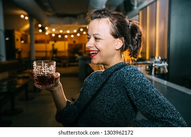 Drunk Brunette Student Woman Alone In Happy Smiling Face Expression Holding And Looking Thoughtful To Scotch Whiskey Glass At Bar Or Pub In Alcohol Abuse And Alcoholic Concept.