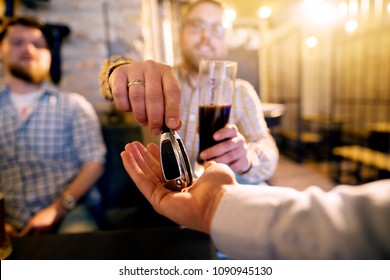Drunk Man With A Beer In Hand Giving Car Key To The Sober Friend While Enjoying In The Bar. Close Up Focus View Of Key And Hands.