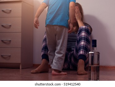 Drunk Mother Sitting On Floor And Covering Her Face From Her Little Scared Son. Child Abuse, Alcoholic Addiction. Glass And Bottle, Shallow Depth Of Field.