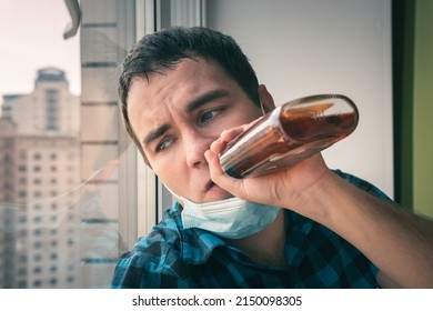 A drunk person is sitting drinks whiskey and looks out window during quarantine of covid 19 coronavirus. Stupid expression of surprise on the drunk's face. A tired employee gets drunk and depressed.