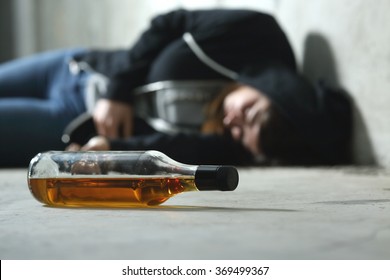 Drunk teenager on the floor in a dark and sad place and an alcohol bottle in the foreground