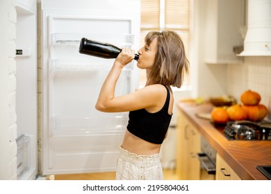 Drunk Woman Drink Some Alcohol From A Bottle, Standing Near Opened Fridge At Home. Alcohol Addiction And Unhealthy Lifestyle Concept