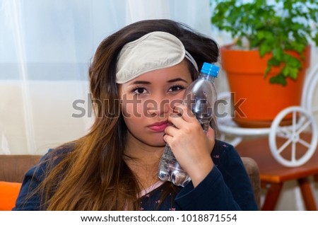 Drunk woman with a sleeping eye mask in her head and with a bottle of water pressing in her face using her other hand, hangover concept