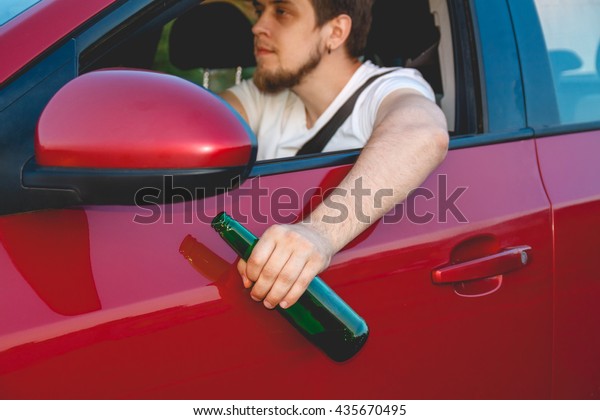 Drunk young man driving a car with a\
bottle of beer. Don\'t drink and drive concept. Driving under the\
influence. DUI, Driving while intoxicated.\
DWI