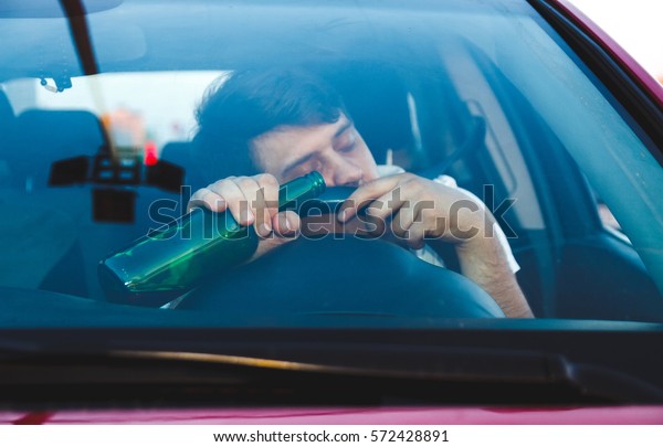Drunk young man driving a car with a\
bottle of beer. Don\'t drink and drive concept. Driving under the\
influence. DUI, Driving while intoxicated.\
DWI