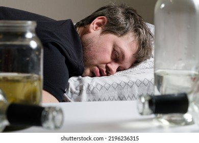 A Drunken Alcoholic Man Lies On The Bed, Empty Bottles On The Table. Alcoholism, Alcohol Dependence As A Social Problem.