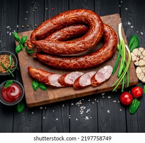 Dry Appetizing Smoked Pork Sausage On A Dark Background With Different Spices And Vegetables. Photo For The Menu