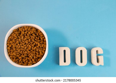 Dry Dog Food In A White Bowl. The Word Dog From White Letters On A Blue Background