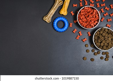 Dry Dog Pet Food In Bowl And Accessories On Blach Chalkboard Background Top View. Pet Feeding Concept Backgrounds With Copy Space. Photograph Taken From Above.