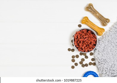 Dry Dog Pet Food In Bowl And Accessories On White Wooden Background Top View. Pet Feeding And Care Concept Backgrounds With Copy Space. Photograph Taken From Above.