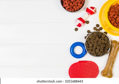 Dry Dog Pet Food In Bowl And Accessories On White Wooden Background Top View. Pet Feeding And Care Concept Backgrounds With Copy Space. Photograph Taken From Above.