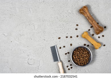 Dry Dog Pet Food In Bowl And Accessories On Gray Concrete Background Top View. Pet Feeding And Care Concept Background With Copy Space. Photograph Taken From Above.