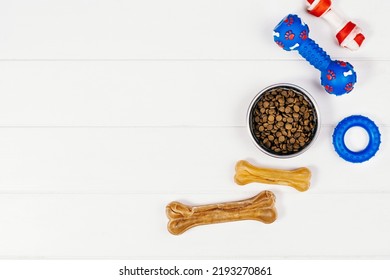 Dry Dog Pet Food In Bowl And Accessories On White Wooden Background Top View. Pet Feeding And Care Concept Background With Copy Space. Photograph Taken From Above.