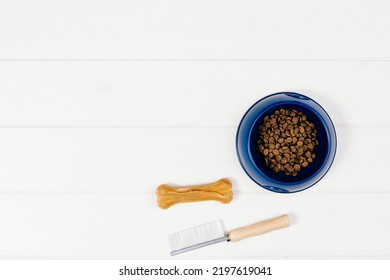 Dry Dog Pet Food In Bowl And Accessories On White Wooden Background Top View. Pet Feeding And Care Concept Background With Copy Space. Photograph Taken From Above.