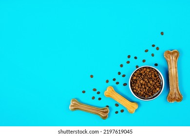 Dry Dog Pet Food In Bowl And Accessories On Blue Background Top View. Pet Feeding And Care Concept Background With Copy Space. Photograph Taken From Above.