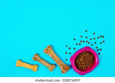 Dry Dog Pet Food In Bowl And Accessories On Blue Background Top View. Pet Feeding And Care Concept Background With Copy Space. Photograph Taken From Above.