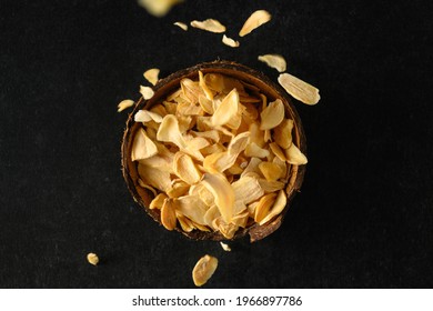 Dry Flakes Of Garlic In A Coconut Bowl, Flakes Falling From Above On A Dark Gray Background, Top View