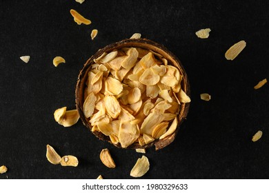 Dry Flakes Of Garlic In A Coconut Bowl, Flakes Falling From Above On A Dark Gray Background, Top View