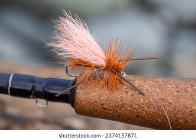 Dry fly pattern in orange with rubber legs on the cork grip of a fly fishing rod at the river.