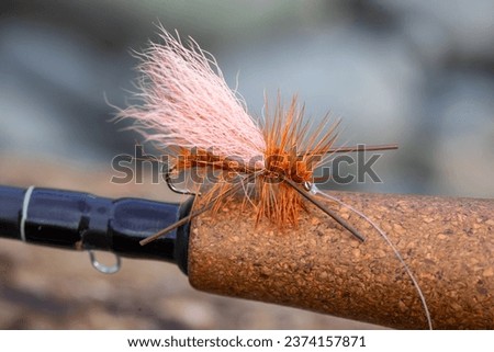 Dry fly pattern in orange with rubber legs on the cork grip of a fly fishing rod at the river.