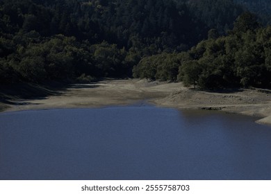 A dry lake offers a serene view, with calm blue water reflecting the surrounding lush greenery. Trees line the banks under a clear sky, showcasing the tranquil beauty of nature in the mountains. - Powered by Shutterstock