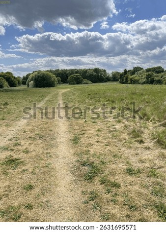 A dry, lightly worn path runs straight down the middle of the field bordered by patchy grass. The ground is slightly yellowed due to dry weather sky is partly cloudy with a dramatic contrast.