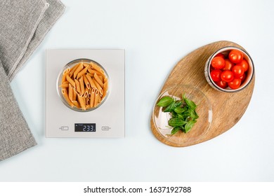 Dry penne pasta in glass bowl on gray digital kitchen scales. Near bowl with red cherry tomatoes and plate with fresh green basil on wooden board. Flatlay. Italian cuisine. Kitchen equipment concept.