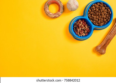 Dry Pet Food In Bowl With A Ball And Dog Bone On Yellow Background Top View