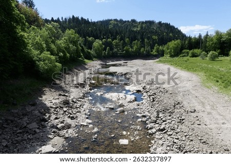 A dry, rocky riverbed winds through lush green forested hills under a bright blue sky, conveying a harmonious natural setting showing climatechange.
