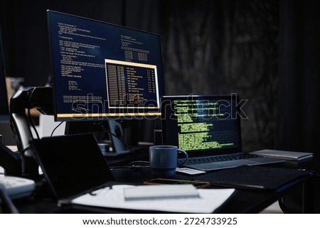 Dual computer monitors displaying programming code on dark screens, laptop and desktop setup on desk with coffee mug, smartphone, and notepad, modern workspace for software development