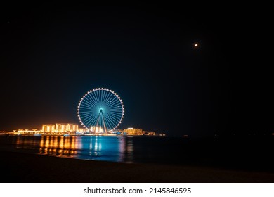 Dubai, UAE - April 2, 2022: Long exposure landscape shot of Ain Dubai ferris wheel and Bluewaters district. Night illumination reflecting on sea surface.