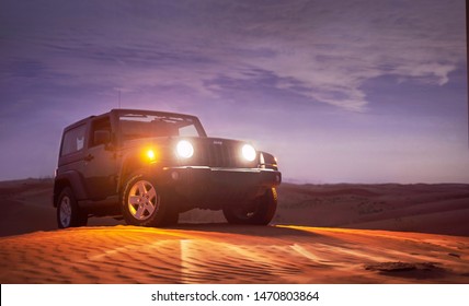 Dubai, UAE - APRIL 23, 2017: Jeep Wrangler Climbing A Sand Dune With The Light On After The Sunset Near Dubai